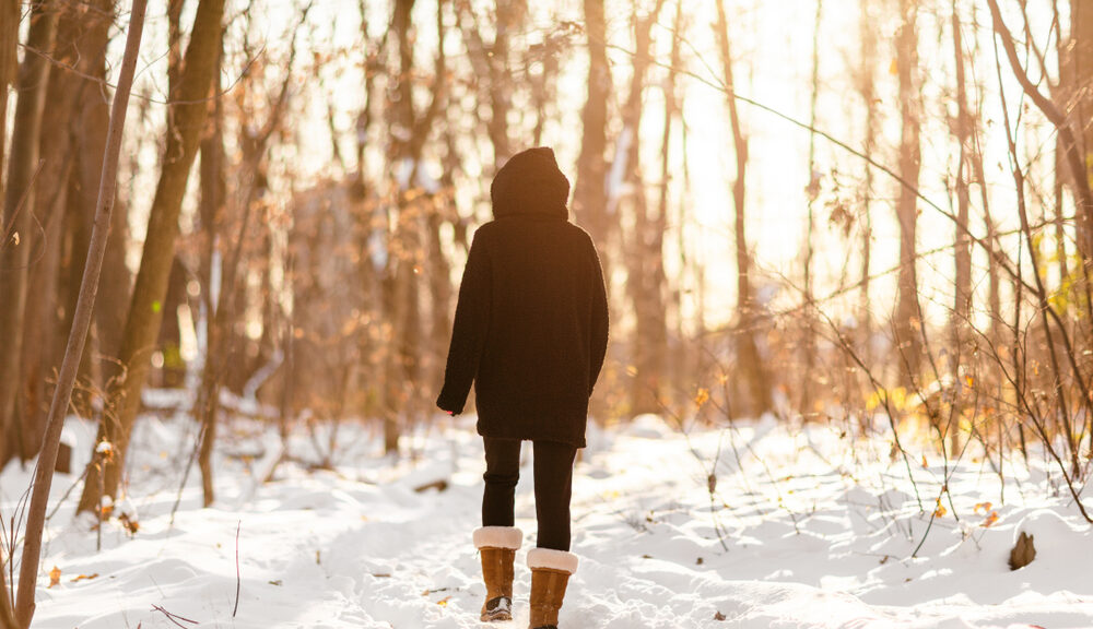 A woman walks through the woods in the snow.