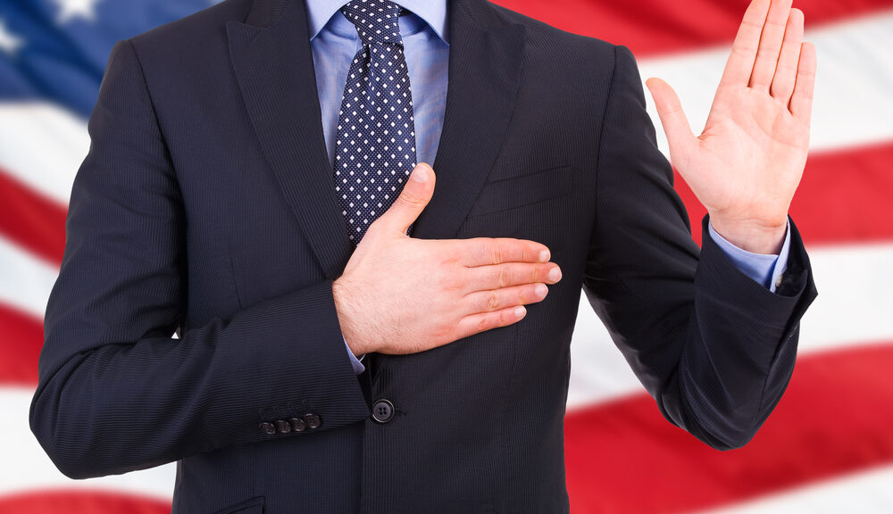 A man holds up his hand and places his hand over his heart with an American flag in the background.