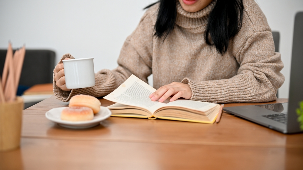 A woman warms her spirit by reading a book, drinking coffee, and eating a biscuit.